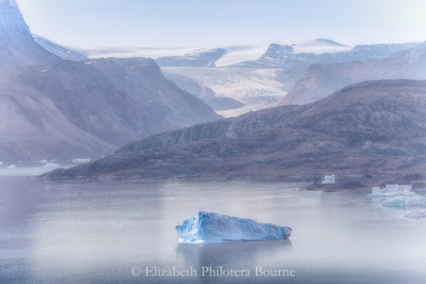 Lone iceberg floating in calm fjord with snow capped mountains and rocky cliffs in Greenland