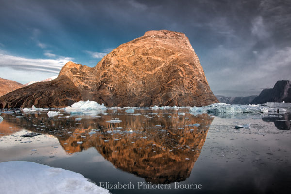 Orange striated rock with dramatic sky reflected in still water with icebergs in Greenland
