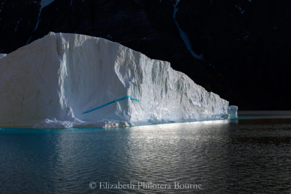 White iceberg floating against black cliff with white reflection glowing in the water in Greenland