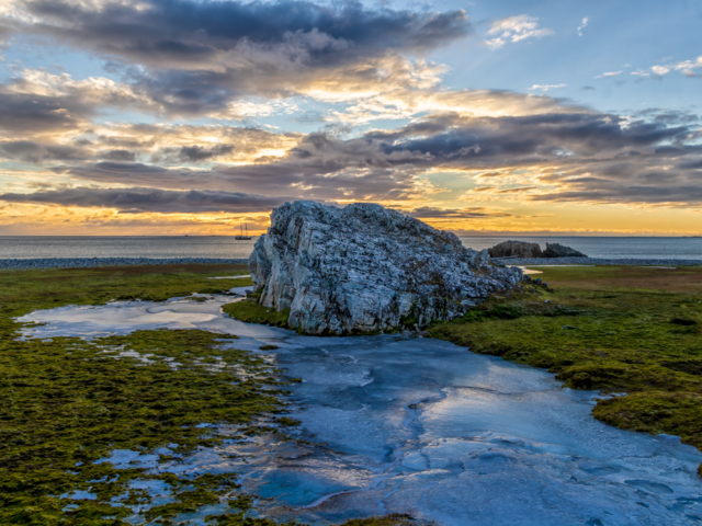 Photograph of a rocks with a frozen stream flowing around it against a sunset sky.