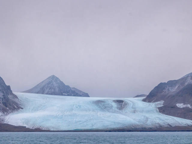Photograph of a soft crevassed glacier with a pale pink sky above.