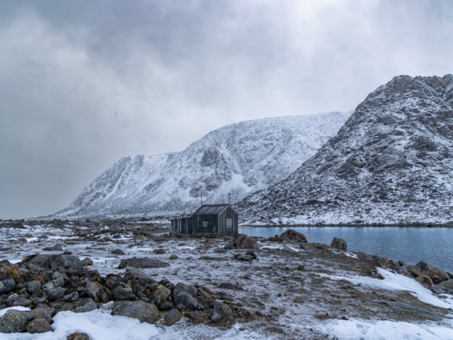 Photograph of a wood and tar paper trappers cabin along a quiet mountain ringed blue bay