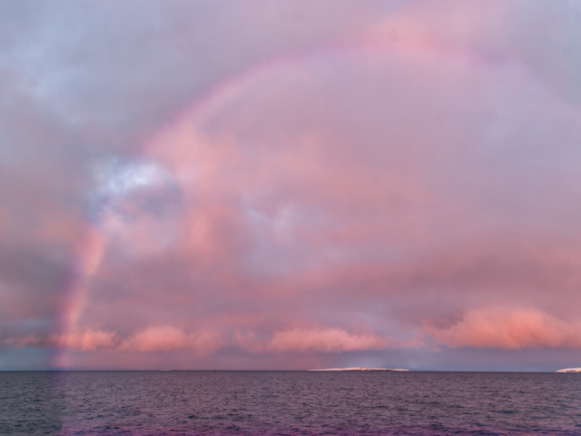 Photograph of a pink sky with a pink rainbow over a slate blue sea
