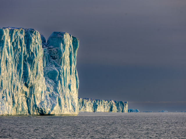 Photograph of blue and white cliffs of ice gainst a dark grey sky