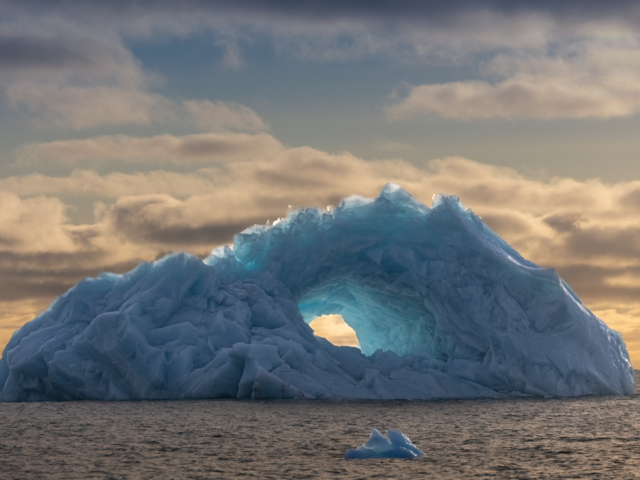 Photograph of a blue iceberg with a hole like an eye in the center against a yellow sky.