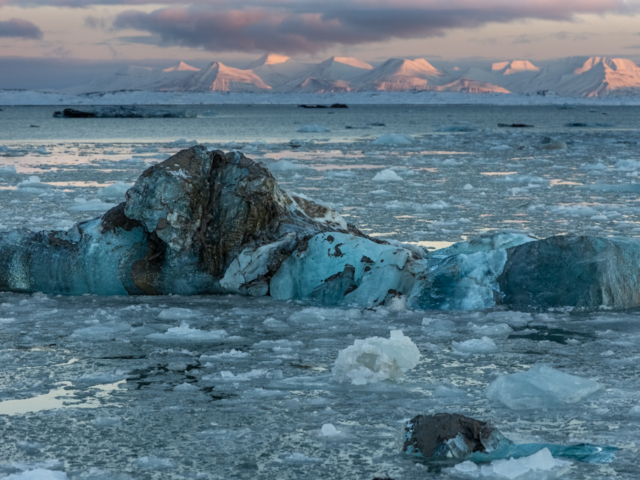 Photograph of a blue and dirty streaked bergy bit with pink mountains in the background.