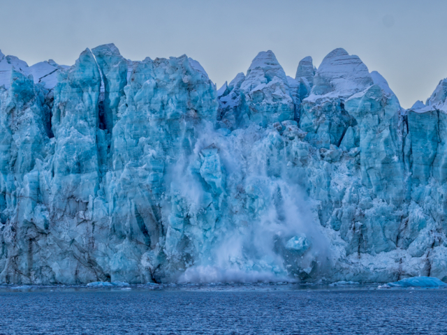 Photograph of a blue glacier with ice falling as the glacier calves.