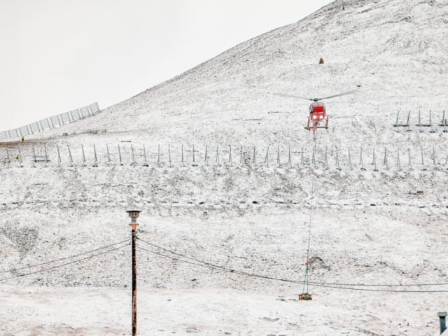 The Avalanche Fence Red helicopter dropping equipment on a snow covered mountain where an avalanche fence is being built